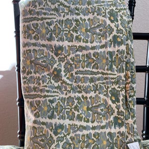 May include: A folded textile with a floral pattern in shades of green, gray, and yellow. The fabric is draped over a black chair, showcasing the intricate design. The pattern features stylized flowers and leaves.