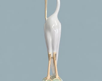 May include: A tall, white ceramic figurine of a standing heron with a long, curved neck. The bird features a gold-painted beak, light tan legs, and stands on a small, circular base with a subtle green wash. The sculpture has a smooth, glossy finish.