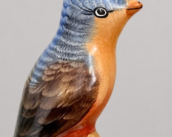 May include: A ceramic bird figurine featuring a blue head with detailed feather markings, a brown back and wings, and an orange chest. The bird has a pointed orange beak and a black eye with painted eyelashes, perched on a light green base.