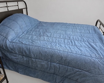 May include: A blue quilted bedspread with horizontal stitching lines covering a bed frame. The bedspread features a textured fabric surface and drapes over the sides of the mattress, providing full coverage for the bed surface in a bedroom setting.