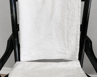 May include: A crisp white textile piece featuring a subtle, tone-on-tone damask pattern with a distinct border. The fabric is folded neatly, showcasing a smooth texture and clean edges, draped over a black wooden chair with a woven seat.