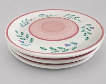 May include: A stack of three round ceramic plates with a white base, featuring a central pink circle and a pink rim. The plates are decorated with a hand-painted pattern of green leafy vines and small blue flowers arranged in a circular motif.
