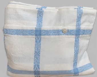 May include: A folded white textile featuring a light blue grid pattern with intricate floral motifs within the stripes. The fabric displays a subtle damask-style weave and includes a single round white button attached to the front edge of the fold.