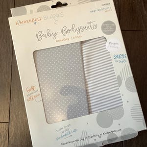 May include: A package of baby bodysuits in Koala Grey, size 6-9 months. The package features two bodysuits, one with white polka dots and the other with gray and white stripes. The packaging includes the text "Soft Premium Cotton!" and "Snuggle in style."