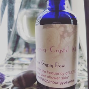 Könnte beinhalten: Eine blaue Glasflasche mit einem weißen Etikett, auf dem "Moon Gypsy Rose Crystal Mist" und "Attune to the frequency of LOVE on your post shower skin*" sowie eine Website-Adresse stehen.