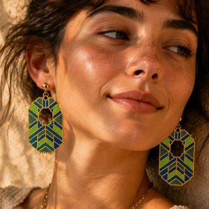 Op de afbeelding: Geometrische kralen oorbellen in groen en blauw, met gouden accenten. De oorbellen hebben een uniek, open middenontwerp. Het model draagt een gouden ketting met een hanger. De oorbellen zijn een statement piece.