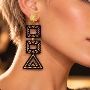 Könnte beinhalten: Ein Ohrhänger mit einem goldfarbenen quadratischen Stecker mit Strahlenstruktur, der mit drei hängenden geometrischen Formen verbunden ist. Diese umfassen zwei schwarze Quadrate und ein schwarzes Dreieck, verziert mit violetten und goldenen Perlenmustern.