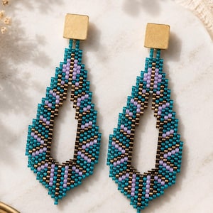 May include: A pair of teardrop-shaped earrings featuring intricate beadwork in shades of teal, lavender, and dark brown. Each earring is topped with a small, square, brushed gold-tone metal stud component that connects to the beaded drop section.