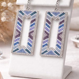 Puede incluir: Pendientes rectangulares de cuentas en tonos azules, morados y turquesas, expuestos sobre un soporte blanco. Los pendientes tienen un patrón geométrico y están suspendidos de ganchos plateados. El fondo es borroso, con tonos blancos y beige.