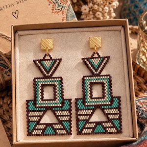 Può includere: Un paio di orecchini geometrici pendenti con un motivo di perline nei toni del turchese, marrone scuro e crema. Gli orecchini hanno un perno quadrato color oro e sono presentati in una scatola con scritto Patrizia Bijoux e un cartellino Good Things Handmade.