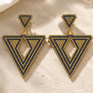 Può includere: Un paio di orecchini geometrici con un design a triangolo. Gli orecchini presentano un metallo color oro con un motivo di perline blu navy e oro. Gli orecchini sono esposti su una superficie chiara.