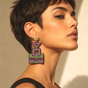 Può includere: Orecchini geometrici di perline nei toni del rosa, verde e nero. Gli orecchini hanno un design a strati con un perno dorato. La modella indossa un top nero.