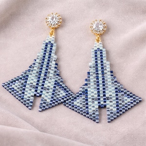 Può includere: Un paio di orecchini di perline fatti a mano. Ogni orecchino presenta un design geometrico nelle tonalità del blu e del bianco, con un perno color oro sormontato da un cristallo rotondo e trasparente. Gli orecchini hanno una forma triangolare.