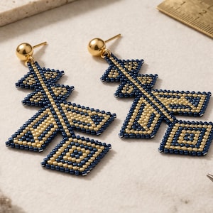 Può includere: Un paio di orecchini pendenti fatti a mano. Gli orecchini presentano un design geometrico con perline blu navy e oro. Ogni orecchino ha un perno a sfera color oro e una forma decorativa. Un righello e delle pinze sono sullo sfondo.