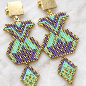 Può includere: Un paio di orecchini pendenti con un motivo geometrico di perline viola e verde chiaro. Ogni orecchino presenta un perno quadrato color oro e un componente di perline a forma di rombo attaccato a una sezione esagonale più grande.