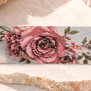 Può includere: Un ampio bracciale di perline con un dettagliato motivo floreale di rose rosa e foglie verdi su sfondo azzurro. Il bracciale è rifinito con terminali in metallo dorato, una chiusura a moschettone e una catenella di estensione regolabile.