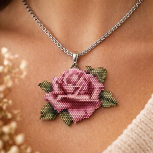 Puede incluir: Un collar con un colgante de rosa de cuentas. El colgante es una rosa rosa y verde detallada con una cadena plateada. La rosa está hecha de pequeñas cuentas, creando una apariencia texturizada. El collar se lleva puesto.
