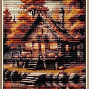 Può includere: Un motivo a punto croce raffigura una scena di un accogliente chalet. Lo chalet, con un tetto scuro e un'illuminazione interna calda, si trova vicino a uno specchio d'acqua riflettente. Intorno allo chalet ci sono alberi con un vivace fogliame arancione e marrone.