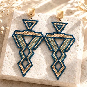 May include: A pair of long, geometric dangle earrings crafted from small, square beads in shades of deep navy blue, metallic gold, and soft sage green. Each earring features a pyramid top connected to a larger, intricate, open-work triangular frame.