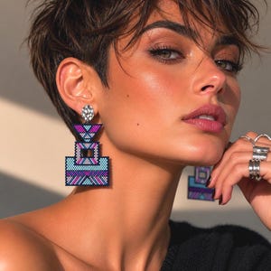 Può includere: Orecchini geometrici di perline nei toni del blu, viola e bianco. Gli orecchini hanno una forma quadrata con un design a strati. La parte superiore dell'orecchino è un perno d'argento. La persona indossa diversi anelli d'argento.