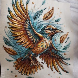 Può includere: Un dettagliato disegno a punto croce di un uccello in volo, con sfumature di oro, marrone e blu. Le ali dell'uccello sono spalancate, circondate da foglie e accenti stilizzati. Il testo "Made by Steph Wilding Design Patrizia Bijoux" è visibile.