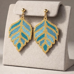 Può includere: Un paio di orecchini a forma di foglia. Gli orecchini sono realizzati con piccole perline color oro e presentano un motivo di perline blu e turchese. Gli orecchini sono esposti su un espositore per gioielli grigio.