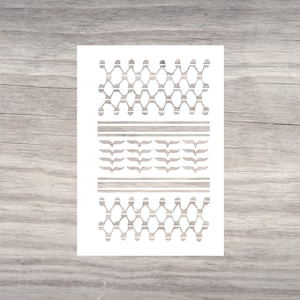 May include: A white stencil with a repeating pattern of geometric shapes and leaf-like designs. The stencil features two rows of connected diamond shapes, two horizontal lines, and a row of stylized leaves. The design is in shades of gray.