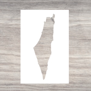 May include: A white stencil cut in the shape of the country of Palestine. The stencil is rectangular and sits on a light gray wooden surface. The stencil is a craft supply for painting or other artistic projects.