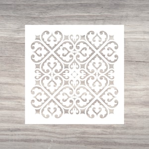 May include: A white stencil featuring a symmetrical, ornate design with swirling, heart-shaped elements. The stencil is square and sits on a light-colored wooden surface, showcasing the intricate pattern.