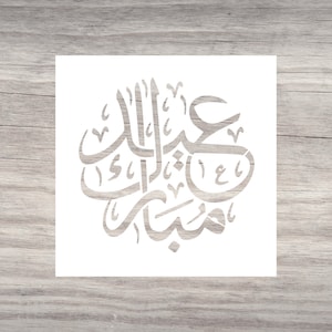 May include: A white stencil featuring Arabic calligraphy, centered on a wooden surface. The calligraphy is in a stylized script, with the words arranged in a circular pattern. The background is a light brown wood grain.