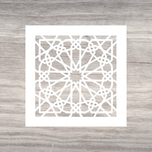 May include: A white geometric stencil featuring an intricate, symmetrical design. The stencil is square and sits on a light gray wooden surface. The design includes star-like shapes and interwoven lines, creating a complex pattern for crafts and decor.