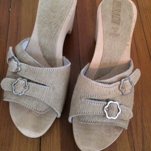 May include: A pair of beige wooden platform sandals with two straps and silver buckles. The sandals have a hairy texture and the brand name "Hush Puppies" is visible on the sole.