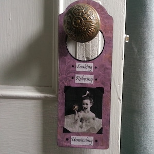May include: A purple door hanger with a brass doorknob. The hanger has a picture of a girl in a bathtub with the words "Soaking", "Relaxing", and "Unwinding" printed on it.