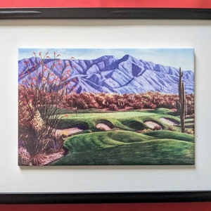 Könnte beinhalten: Ein gerahmter Druck eines Golfplatzes mit einer Bergkette im Hintergrund. Der Golfplatz ist grün und die Berge sind lila. Im Vordergrund befinden sich Bäume und Kakteen.