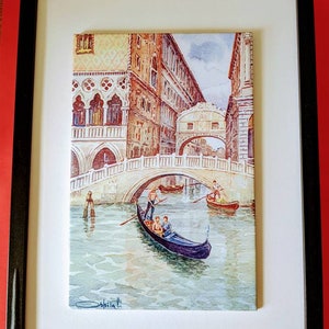 May include: A watercolor painting of a gondola in a canal in Venice, Italy. The gondola is being rowed by a gondolier and there are passengers in the gondola. The painting is framed in a black frame.