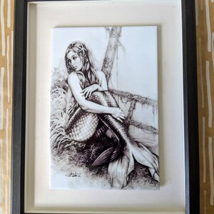 Peut inclure: Un dessin encadré en noir et blanc d'une sirène assise sur un rocher avec sa queue dans l'eau. Le dessin est sur une tuile blanche et a un cadre noir.