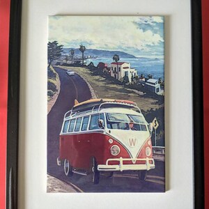 May include: A framed vintage postcard featuring a red and white Volkswagen bus driving along a coastal road with houses and palm trees in the background.
