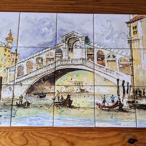 May include: A watercolor painting of a bridge in Venice, Italy. The bridge is white with arches and is over a canal. There are gondolas in the water and buildings in the background.