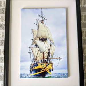Op de afbeelding: Een ingelijste print van een vintage zeilschip met witte zeilen tegen een blauwe lucht. Het schip is geschilderd in tinten blauw, geel en zwart.