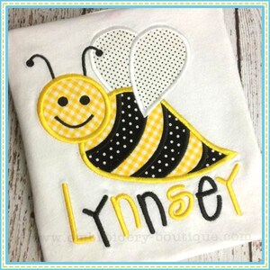 May include: Embroidered design of a yellow and black bee with a gingham pattern on the body and wings. The bee has a smiling face and is holding a yellow flower. The name "Lynsey" is embroidered below the bee.