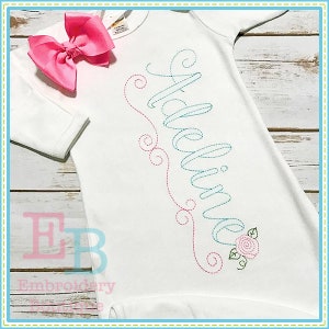 Op de afbeelding: Witte baby romper met de naam "Adelaide" geborduurd in blauw en roze garen met een roze strik op de schouder.