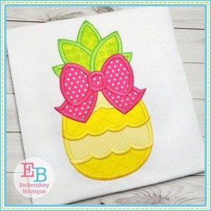 May include: Embroidered design of a yellow pineapple with a pink polka dot bow. The pineapple has green leaves on top. The design is on a white background.