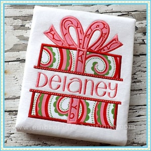 May include: Embroidered design of a red and green gift box with a pink bow and the name "Delaney" on a white t-shirt.