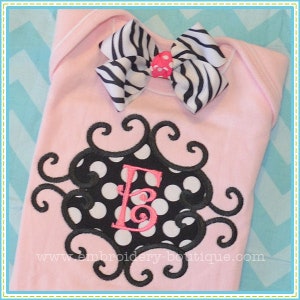 May include: A pink baby onesie with a black and white zebra print bow and a black and white polka dot monogram with a pink letter E in the centre. The monogram is surrounded by a black swirl design.