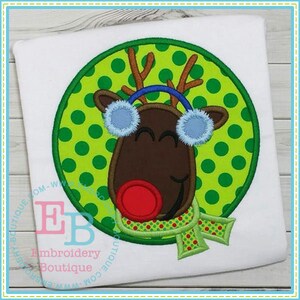 May include: Embroidered applique of a cartoon reindeer wearing a blue headband and earmuffs, a red nose, and a green and red scarf. The reindeer is on a green background with white polka dots.
