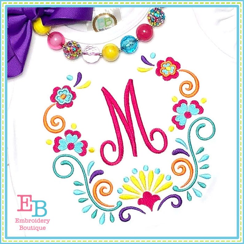 May include: White t-shirt with a colorful embroidered floral design and the letter "M" in the center. The design features swirls, flowers, and leaves in shades of pink, blue, yellow, orange, and purple.