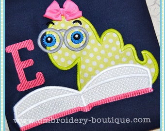 May include: Embroidered design featuring a cartoon worm wearing glasses and a pink bow, reading a book. The worm is yellow with white polka dots. A pink letter "E" is on the left. The website address "embroidery-boutique.com" is at the bottom.