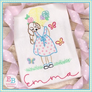 May include: Embroidered design of a girl with a dog, wearing a pink polka dot dress and a flower crown. The design includes butterflies and the name "Emma" in cursive.