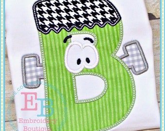 May include: Green letter B with a black and white houndstooth pattern on top, white eyes, and a gray and white checkered arm. The letter is stitched on a white fabric.