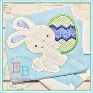 May include: White bunny holding a green, blue and white chevron patterned Easter egg. The bunny has blue ears and a pink nose. The bunny and egg are embroidered on a light blue fabric.